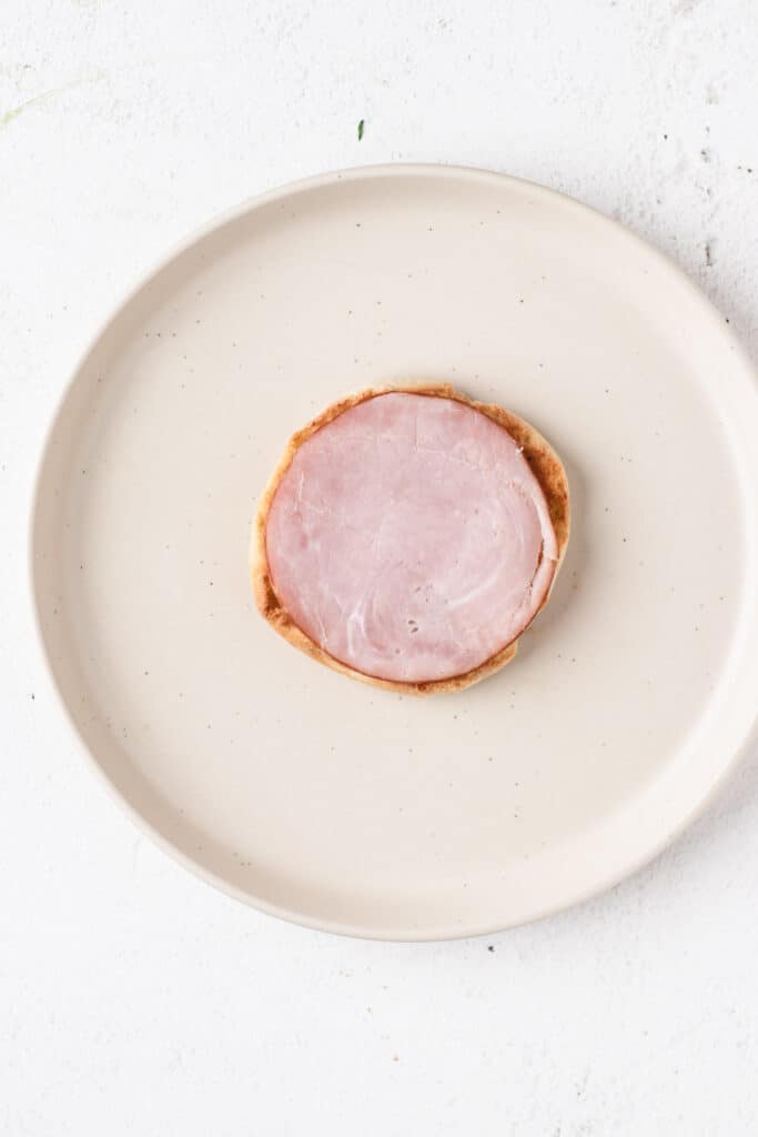 Placing a slice of Canadian bacon on a toasted English muffin.