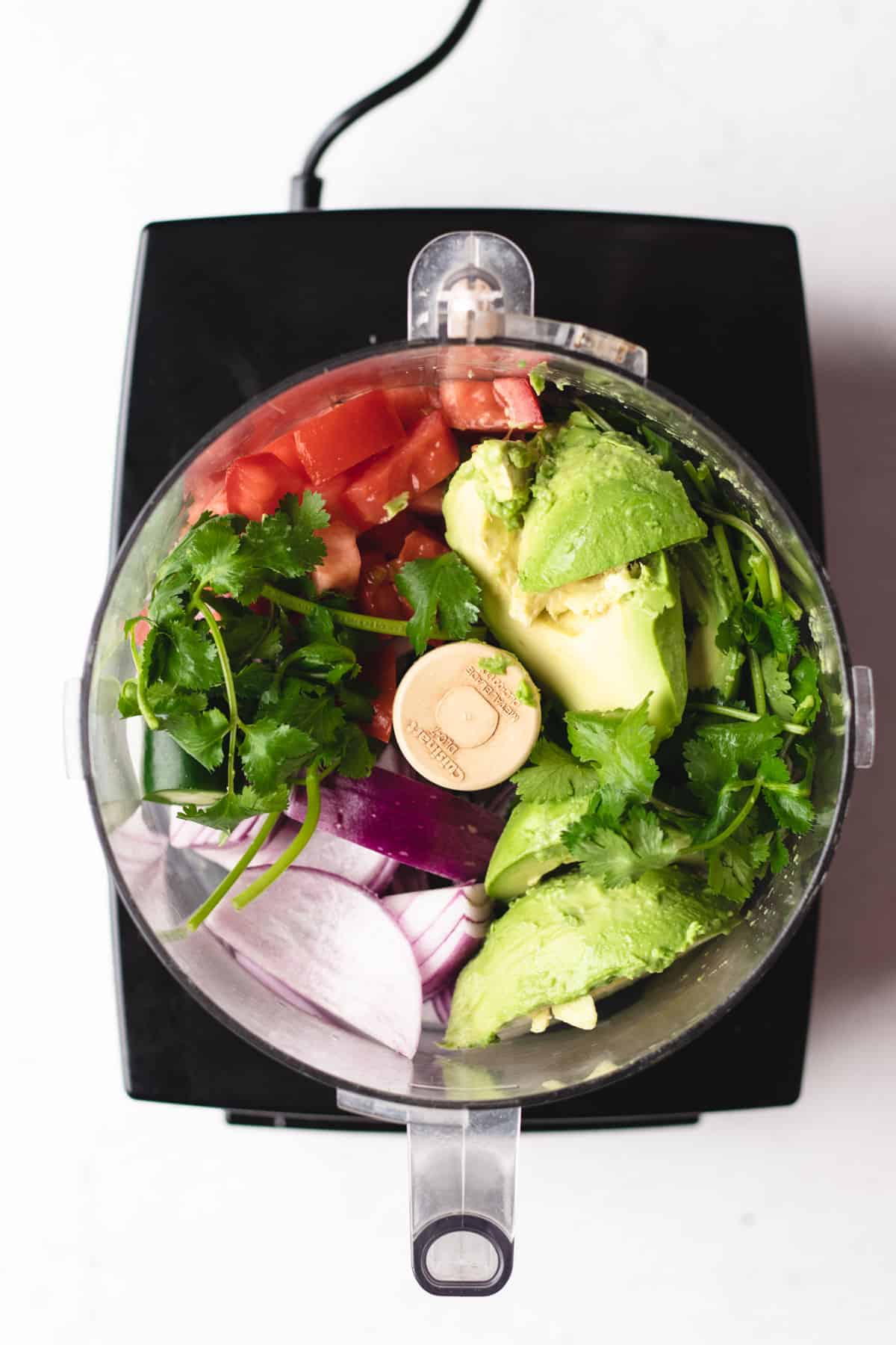 Avocados, tomatoes, onions, and cilantro are blended in a food processor.