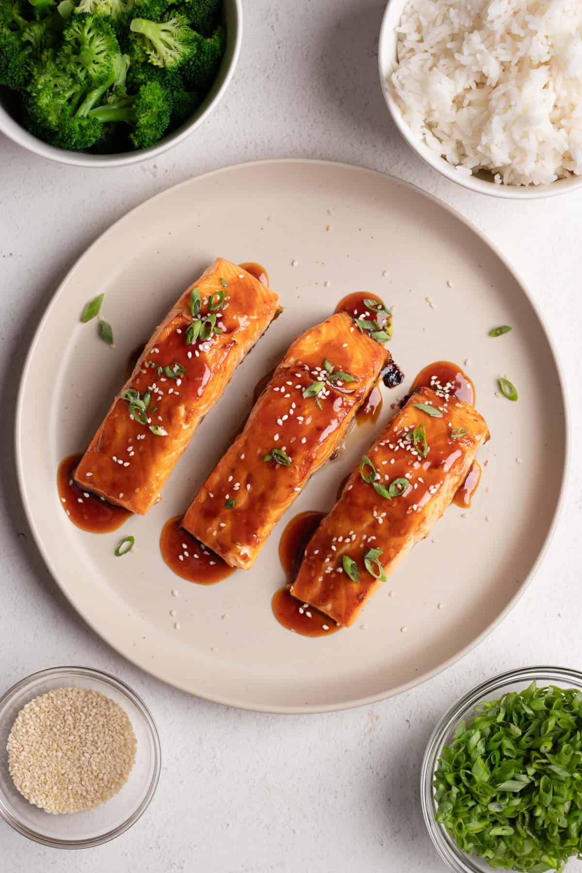 Three pieces of teriyaki salmon on a large round plate with sesame seeds and green onions.