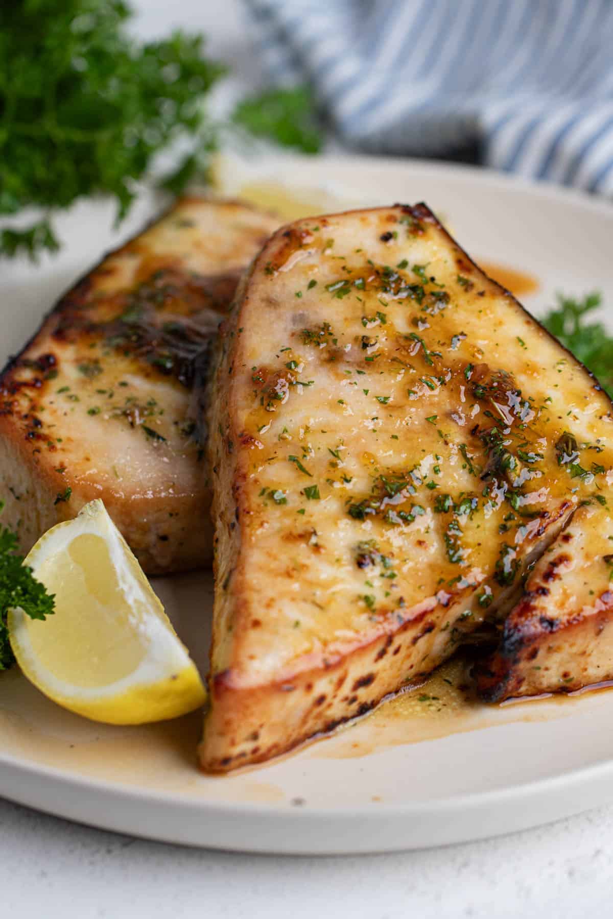 Two swordfish steaks on a large round white plate garnished with parsley and lemon.