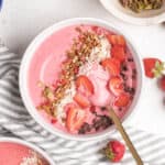 A round bowl with a strawberry smoothie, topping, and a golden spoon.