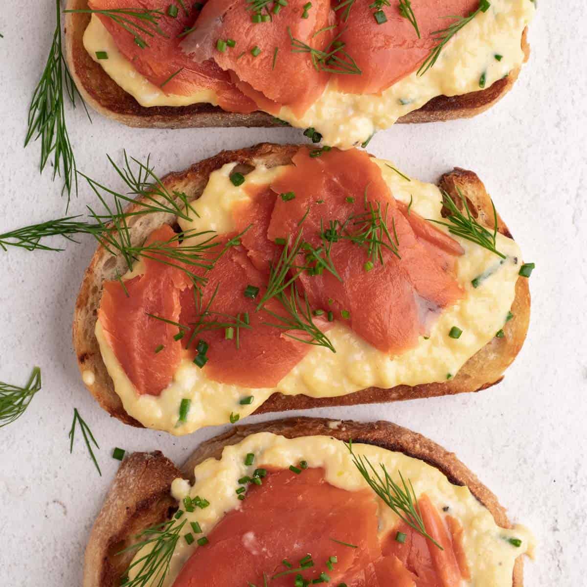 Three slices of toast with scrambled eggs and smoked salmon garnished with fresh dill.