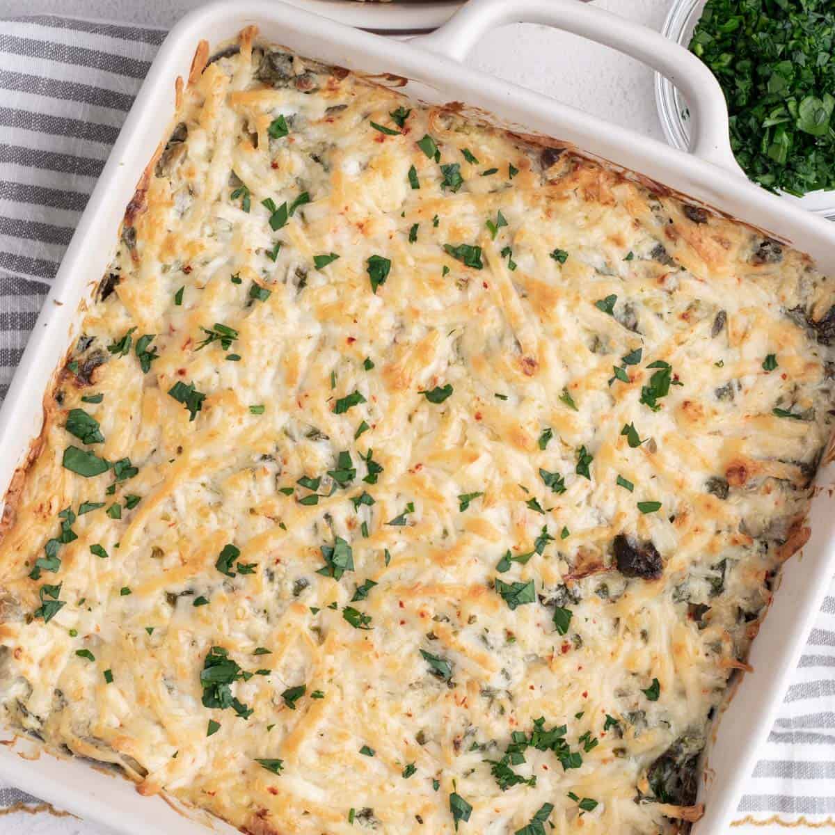 Baked artichoke and spinach dip in a square casserole dish with chopped parsley on top.