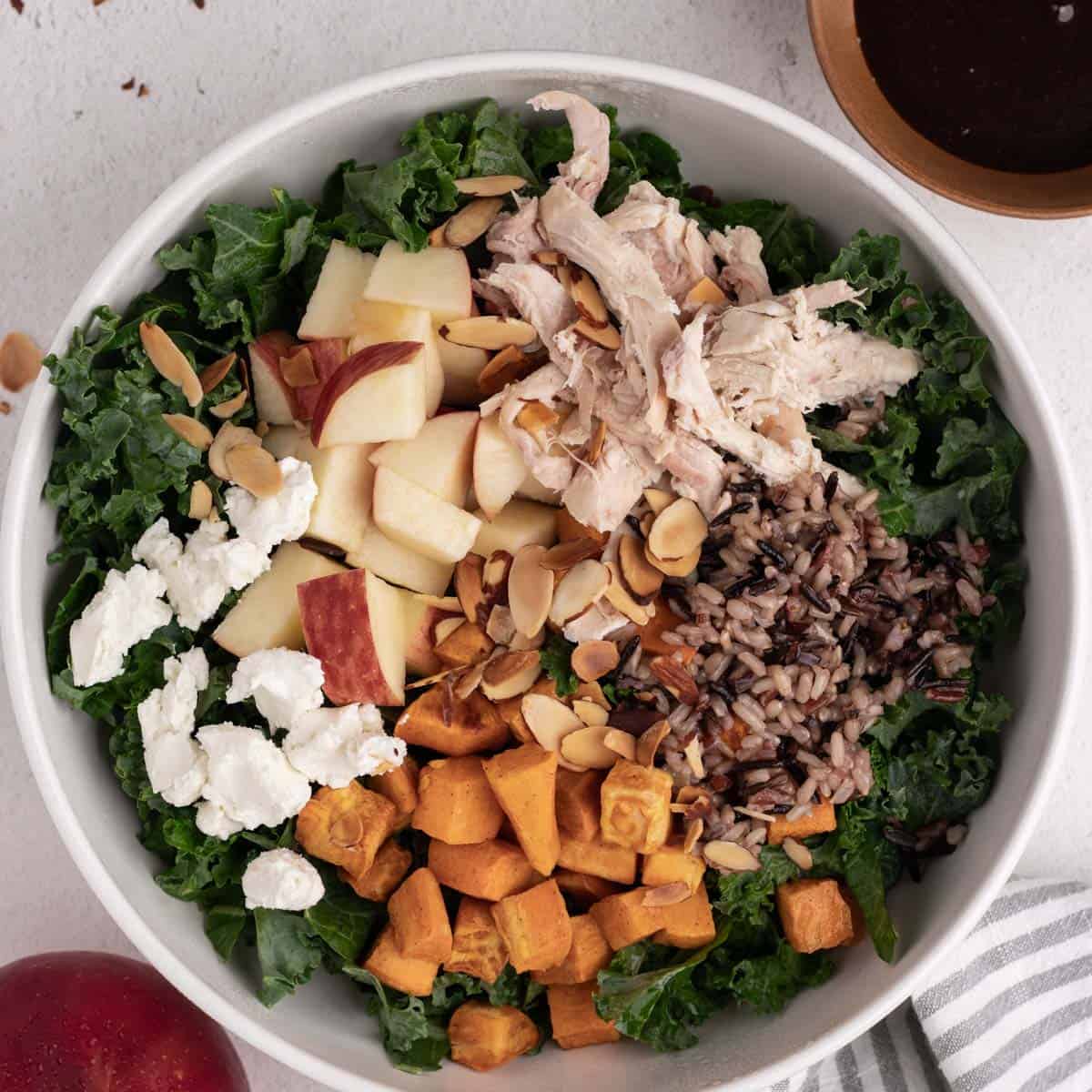 A round white bowl filled with kales, sweet potatoes, almonds, chicken, apples, and goat cheese.