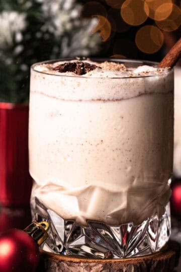 A crystal glass filled with eggnog with Christmas lights in the background.