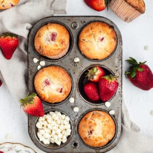 Strawberry muffins, whole strawberries, and white chocolate chips inside a muffin pan.