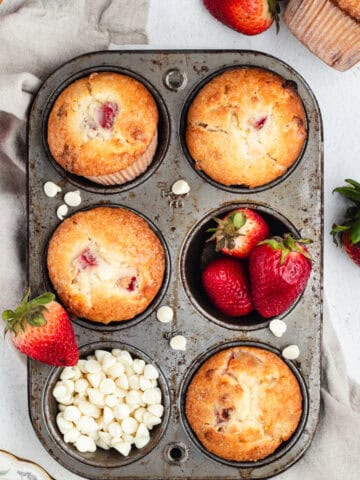 Strawberry muffins, whole strawberries, and white chocolate chips inside a muffin pan.
