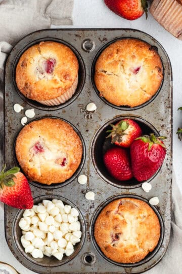 Strawberry muffins, whole strawberries, and white chocolate chips inside a muffin pan.