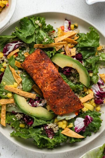 A filet of blackened salmon on top of a salad of leaf lettuce, mango salsa, and sliced avocados.