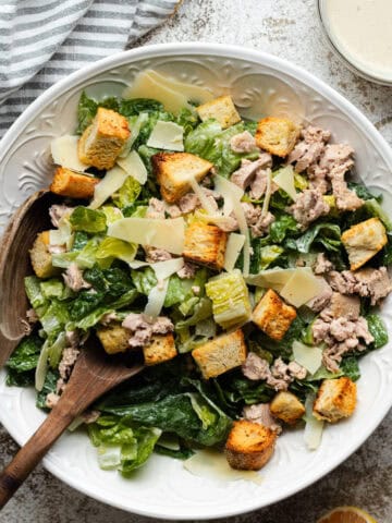 Tuna Caesar salad in a large round white bowl with wooden serving spoons.