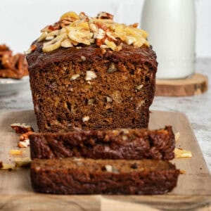 Whole banana bread loaf on a wooden cutting board with two slices placed in front, showing the moist texture and golden-brown crust.