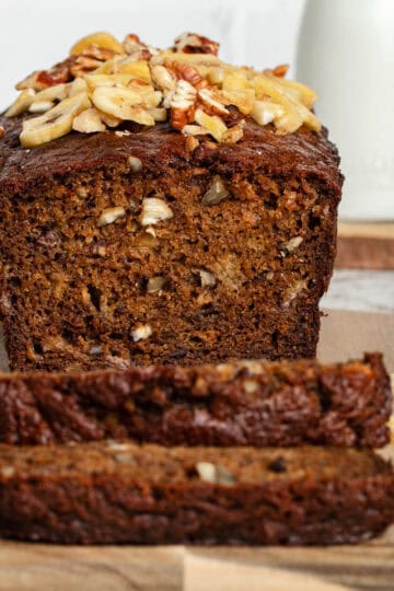Whole banana bread loaf on a wooden cutting board with two slices placed in front, showing the moist texture and golden-brown crust.