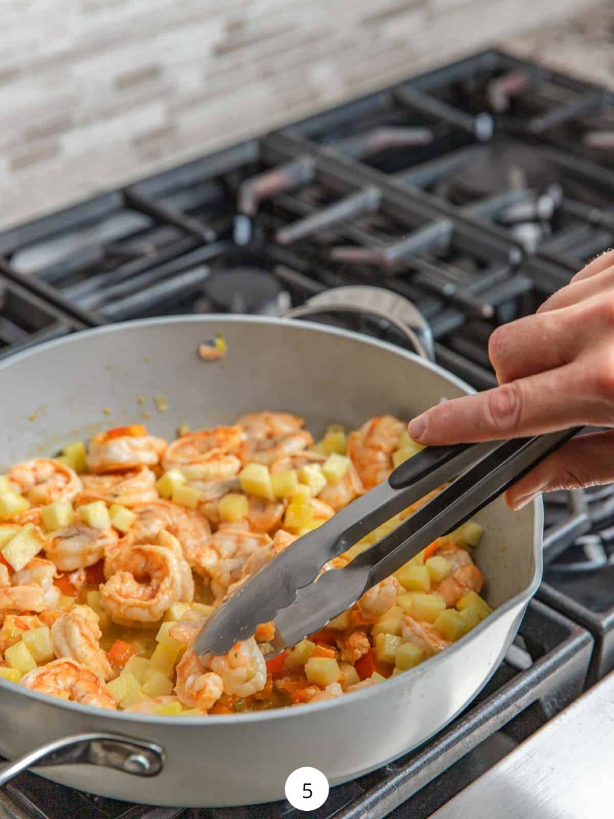 Cooking shrimp in pineapple in a large pan and turning the shrimp over using metal tongs.