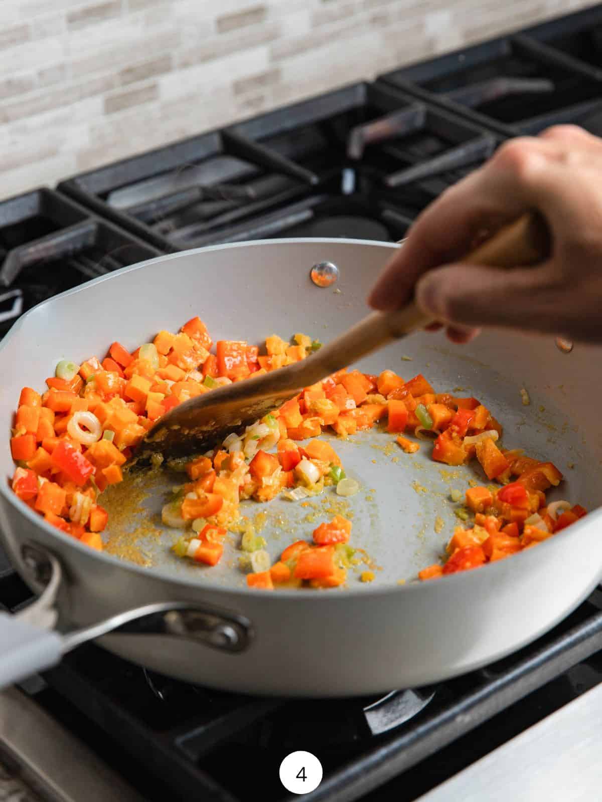 Cooking carrots, and red bell pepper in a large pan using a wooden spoon.