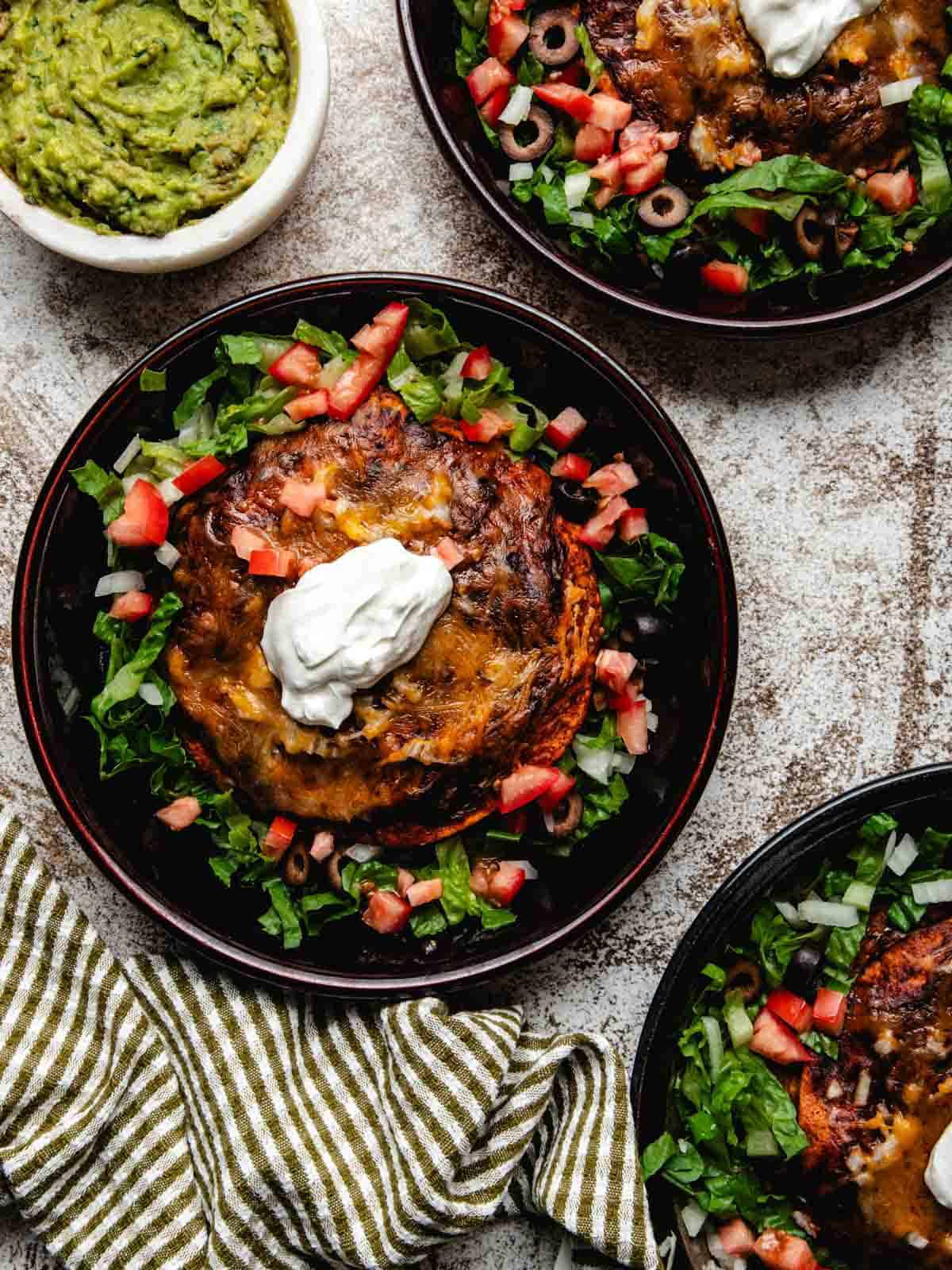 Three shallow bowls of stacked enchiladas with a small bowl of guacamole off to the side.