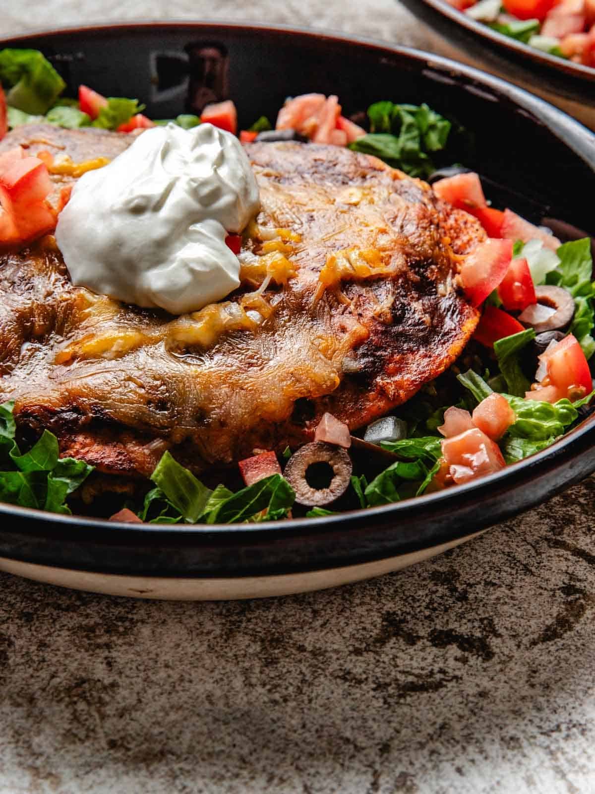 Stacked beef enchiladas inside of a shallow bowl garnished with lettuce, tomato, olives, and sour cream.
