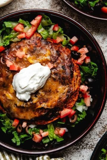 Three shallow bowls of stacked enchiladas garnished with lettuce, tomatoes, olives, and sour cream.