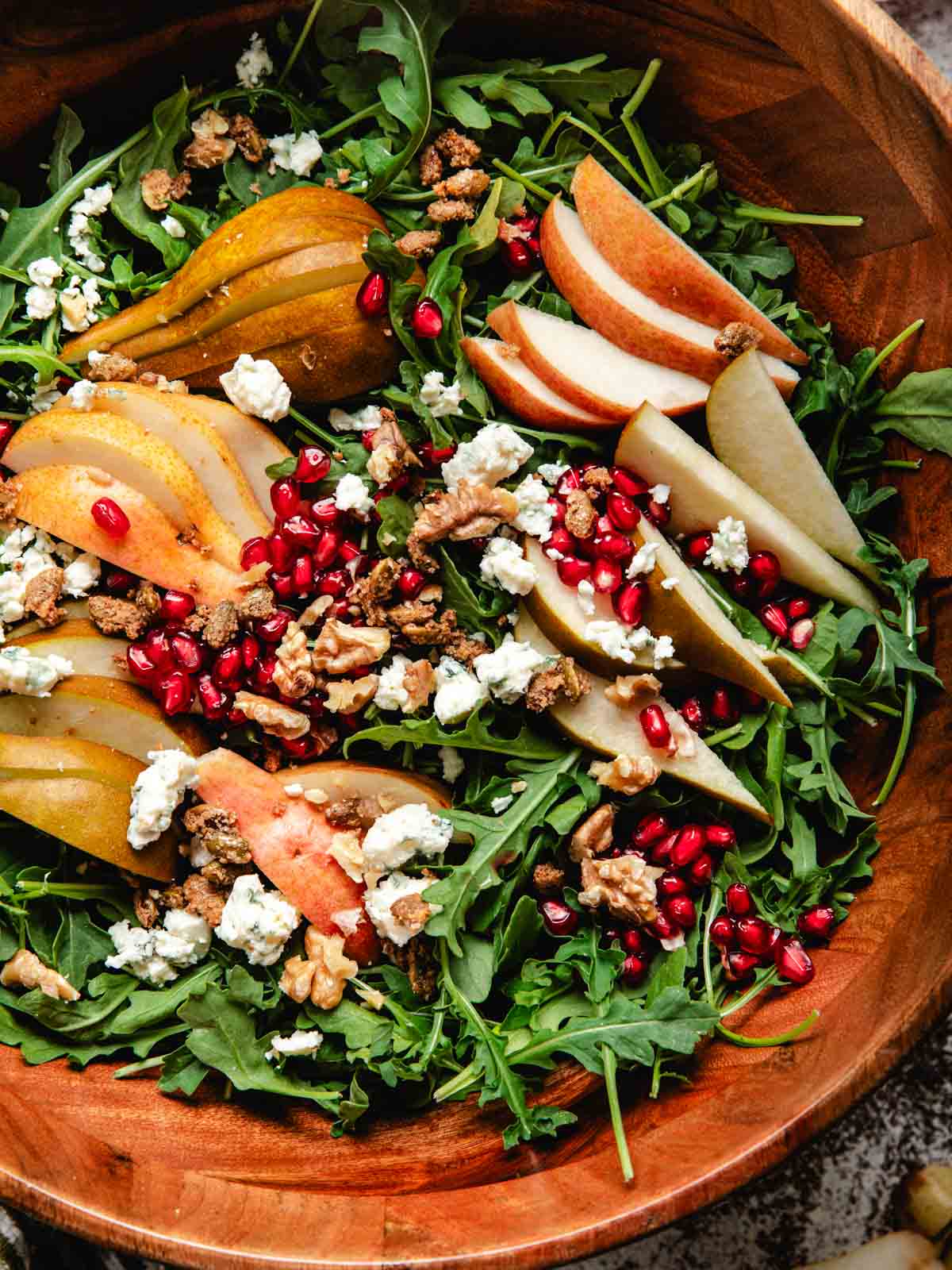 A large wooden salad bowl filled with fresh arugula, sliced pears, candied pepita seeds, walnuts, pomegranate seeds, and crumbled blue cheese.