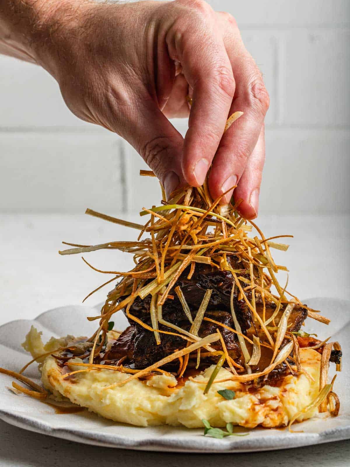 A hand placing frizzled leeks over top of braised short ribs for a garnish.