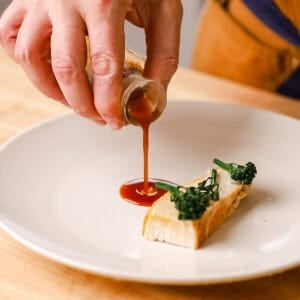 Pouring chicken demi glace in a thin stream onto a white plate with chicken and broccolini.
