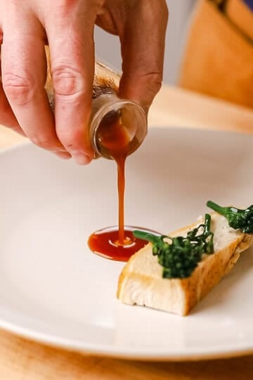 Pouring chicken demi glace in a thin stream onto a white plate with chicken and broccolini.
