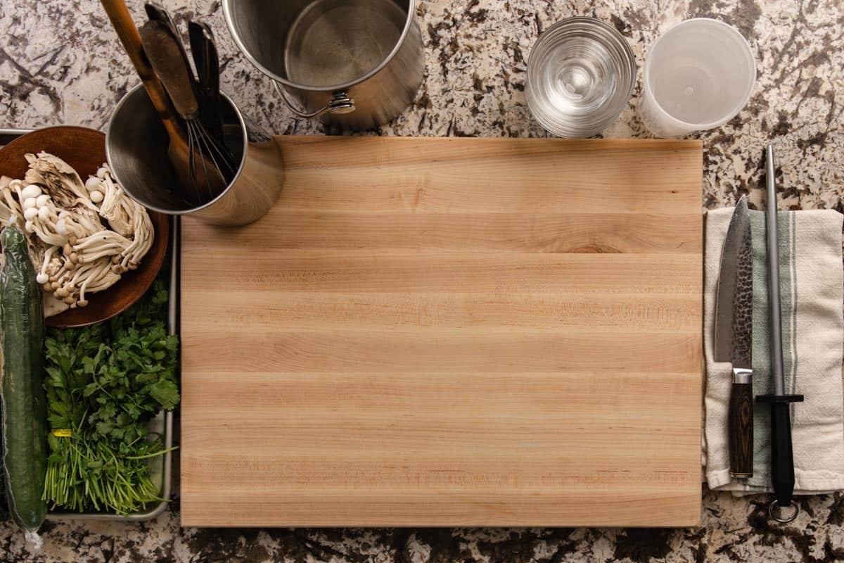 A wooden cutting board, chef knife, honing rod, food waste container, and kitchen utensils.