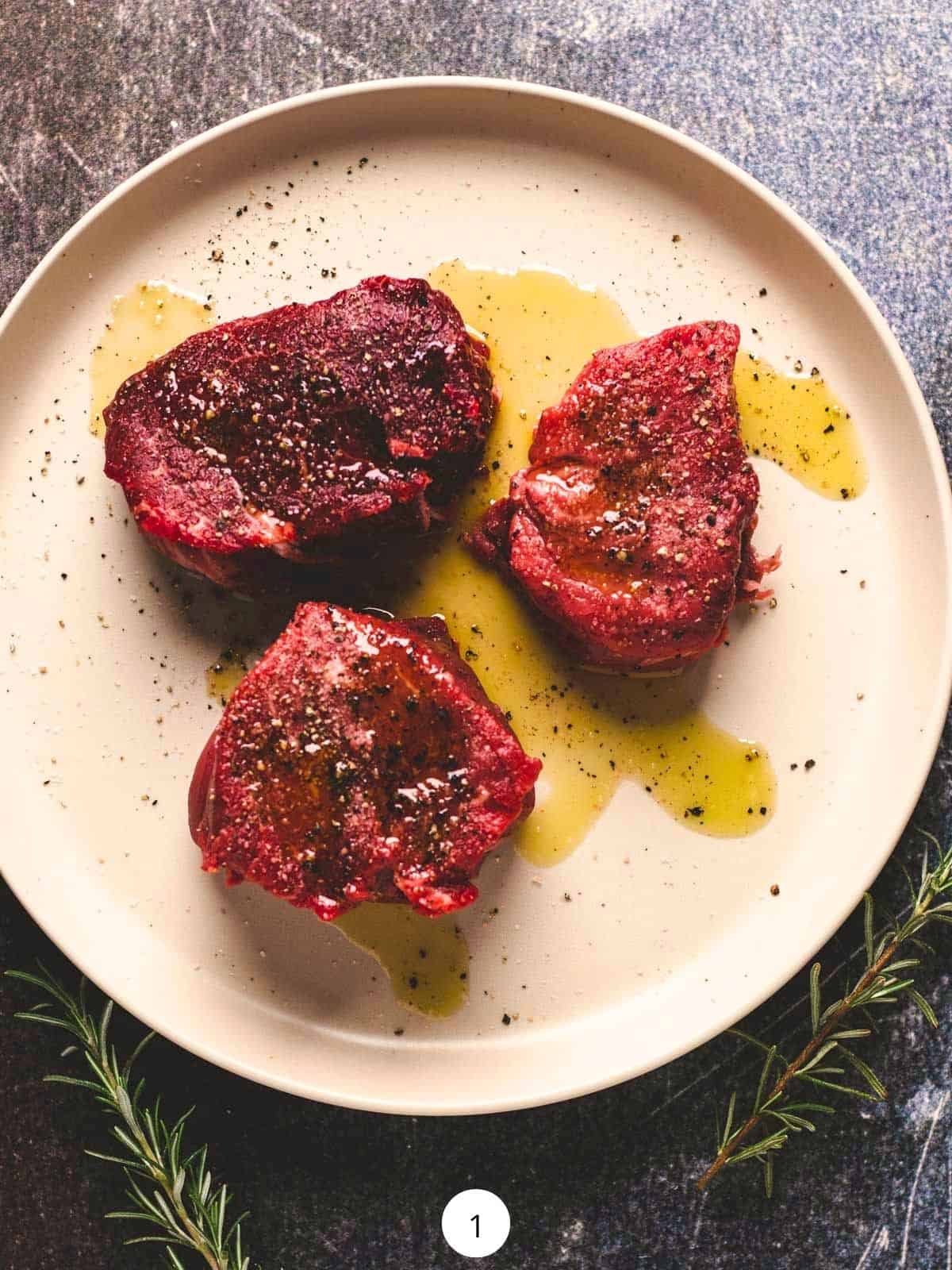 Three filet mignons on a round plate seasoned with olive oil, salt, and pepper.